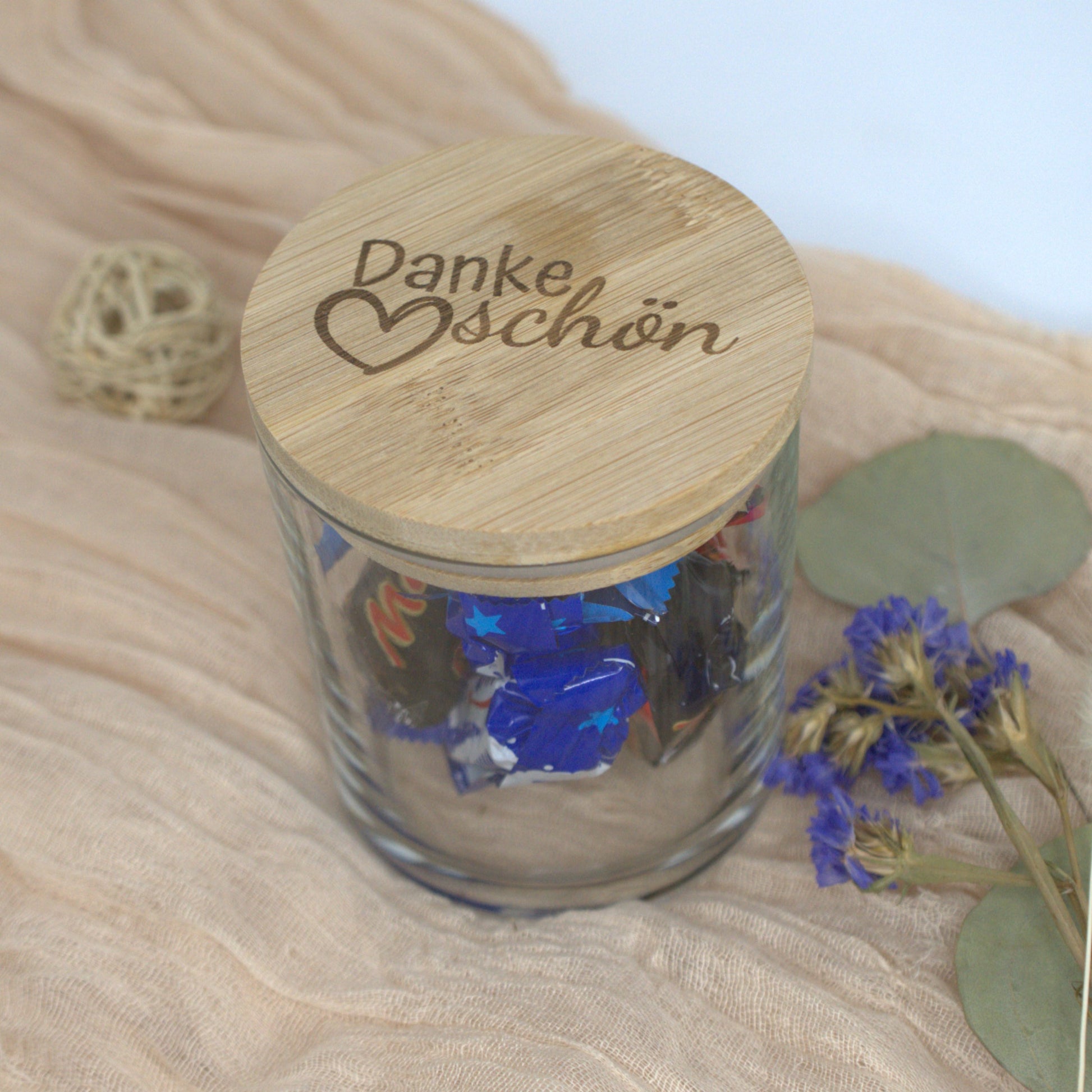 Geschenkglas mit Bambusdeckel und Gravur ‚Dankeschön‘, gefüllt mit Süßigkeiten, auf dekorativer Stoffunterlage